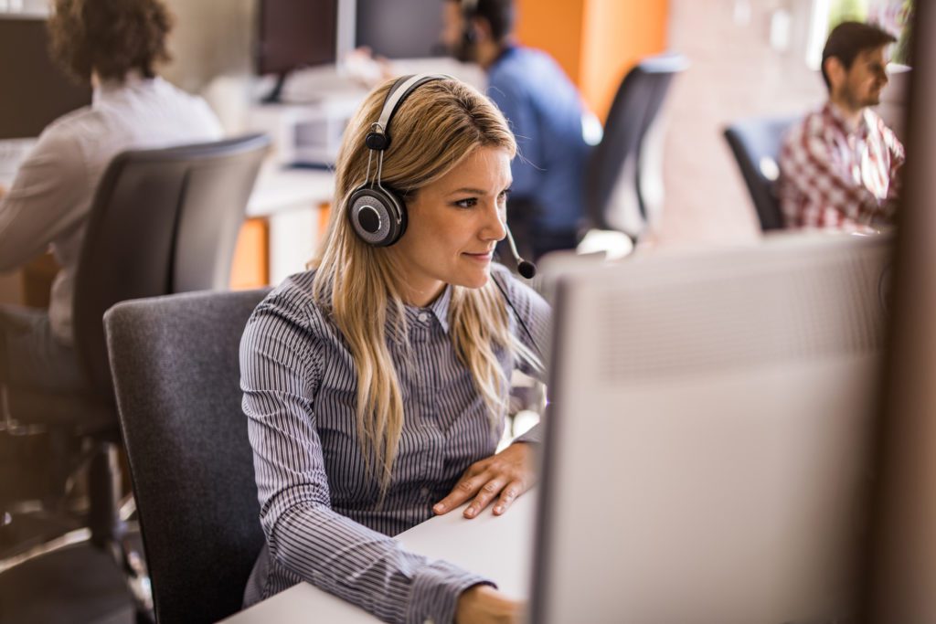 Young woman with headset working on desktop PC at call center. Her colleagues are in the background.