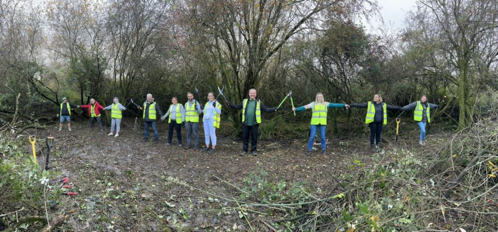 11 adults in high-visibility vests, in a line holding gardening tools in a woodland area